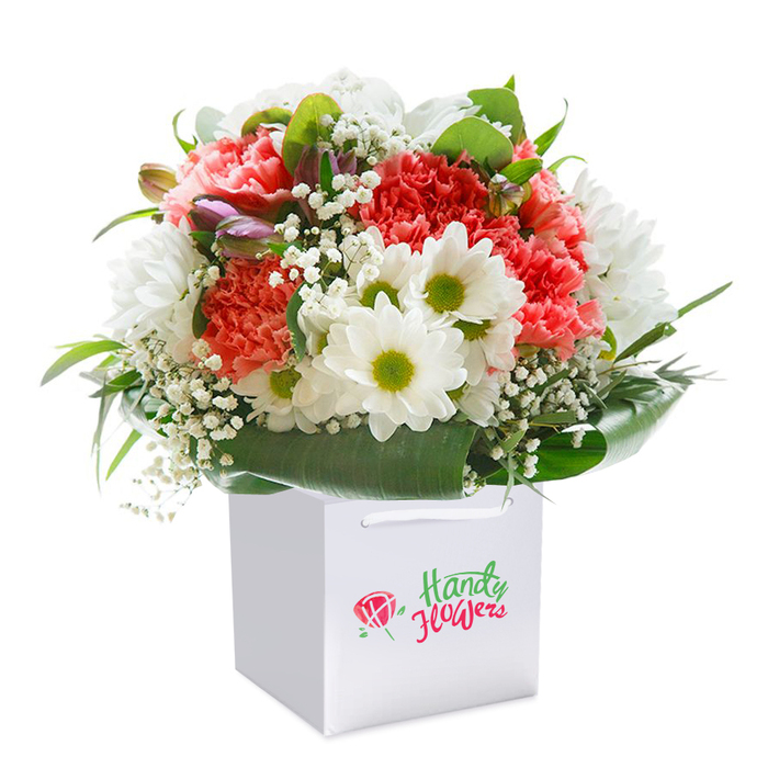 This beautifully layered bouquet, poised within a square white gift bag, evokes both celebration and serenity-a fitting choice for South Ruislip's vibrant yet grounded atmosphere. The star attractions are soft coral-pink and peach carnations, their petals richly ruffled and tenderly arranged for an inviting fullness. Stately white daisies punctuate the bouquet, glowing from their chartreuse-yellow centers, and create a cheerful atmosphere reminiscent of garden scenes near Ruislip Lido or along Whitby Road. Ethereal sprays of baby's breath add subtle lightness, with their dainty, cloud-like florets softening the bolder colours. Discreetly hidden within, purple-tipped buds-possibly shy alstroemeria-appear as whispers of colour, deepening the arrangement's composition and offering an inviting sense of discovery. Glossy, broad green foliage wraps low around the base, completing the design with a touch of natural grandeur. The bouquet is expertly shaped into a full, rounded silhouette, highlighted by balanced, even lighting that showcases each detail upon a simple white canvas. Designed for easy carrying and elegant gifting, the sturdy white-corded handles speak of both convenience and style, perfect for same-day delivery to homes and businesses throughout the HA4 area. Each element and flourish reflects the experienced touch of South Ruislip's florists, making this arrangement a heartfelt expression for any joyful or meaningful occasion.